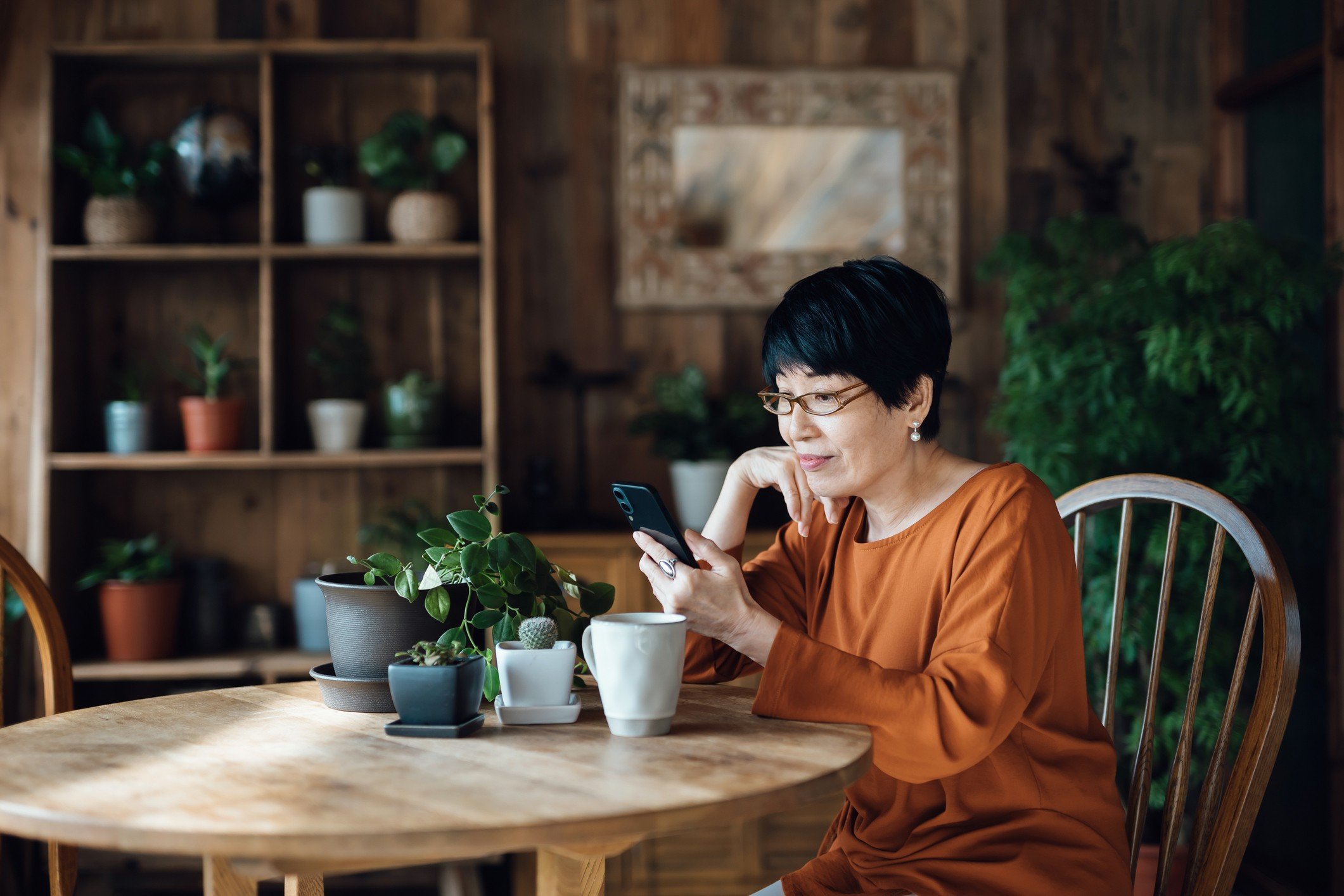 Middle-aged woman with glasses smiles while using her smartphone to browse peer-to-peer lending loans in a cozy, plant-filled room.