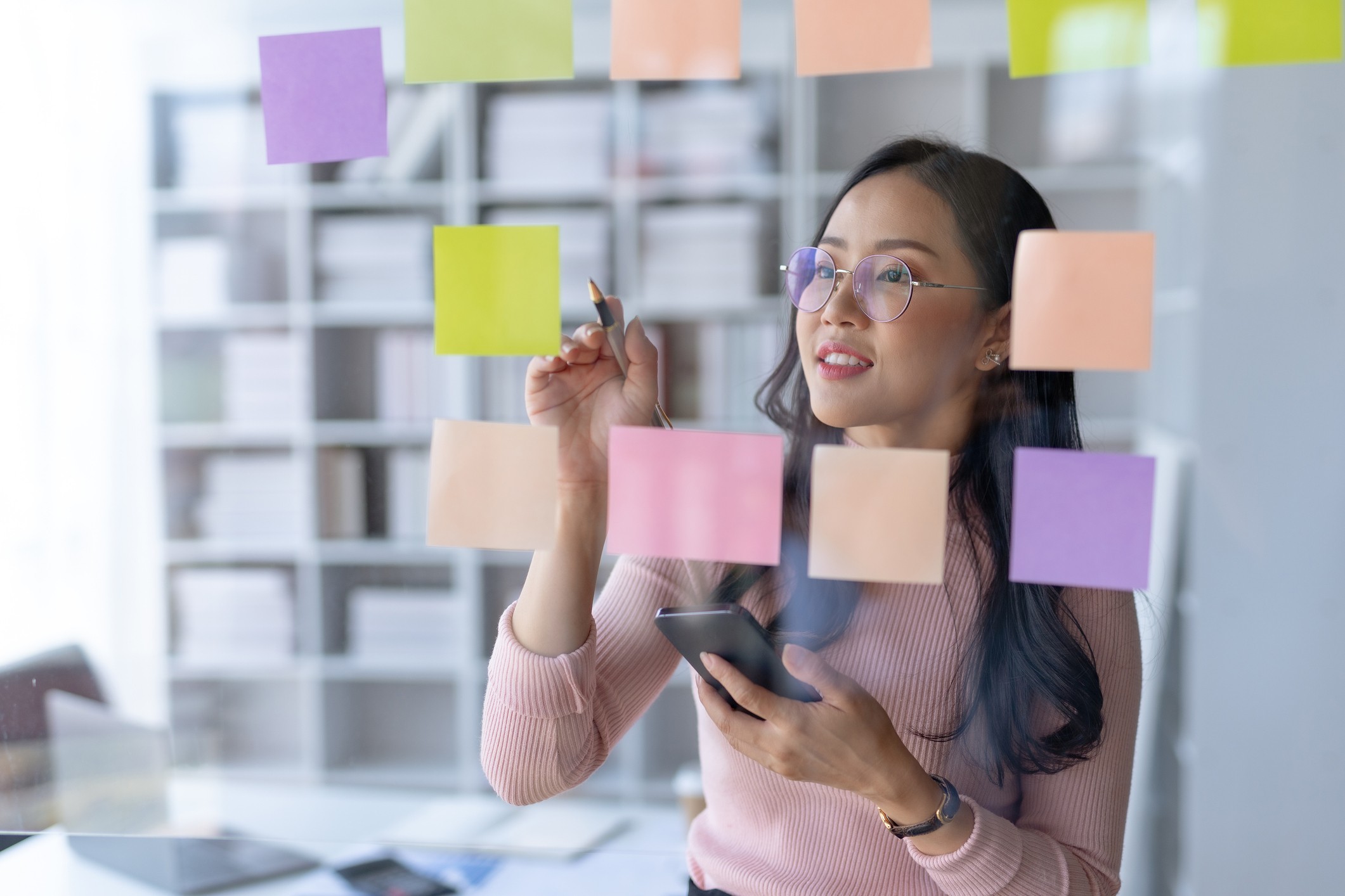 Woman creating a plan on sticky notes, considering strategies for paying off debt while using her smartphone as a reference