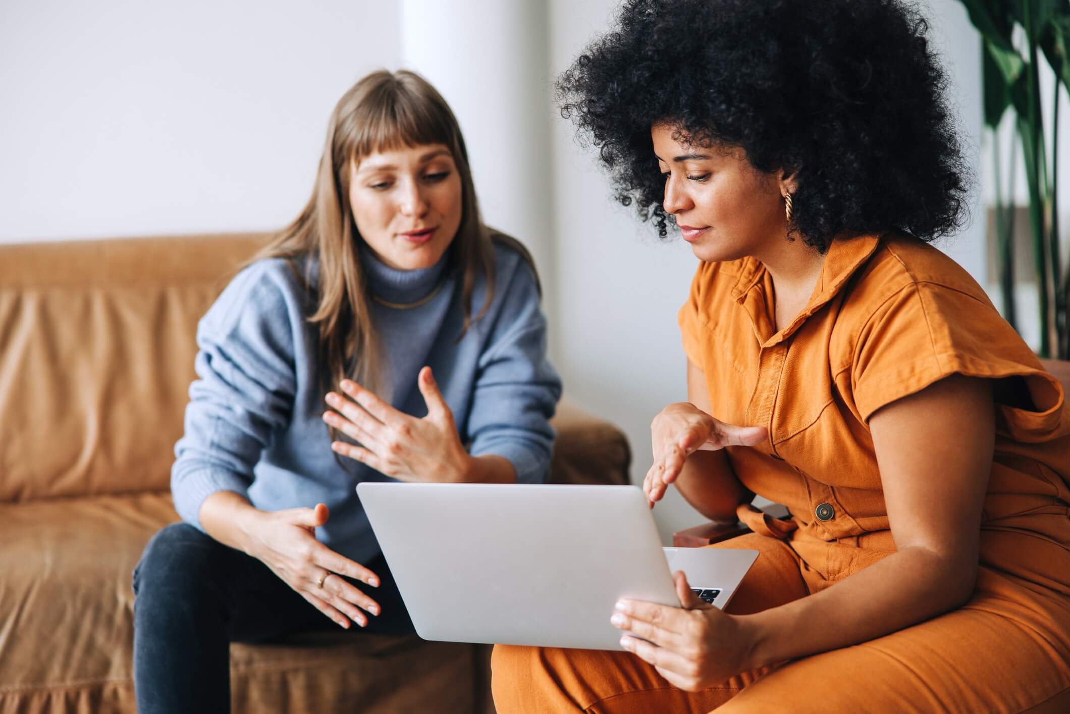 Two women are sitting on a couch, discussing finances while looking at a laptop, possibly exploring options like using a personal loan to pay taxes.