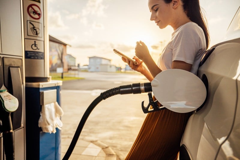 young woman text messaging on smart phone while refueling gas tank at fuel pump