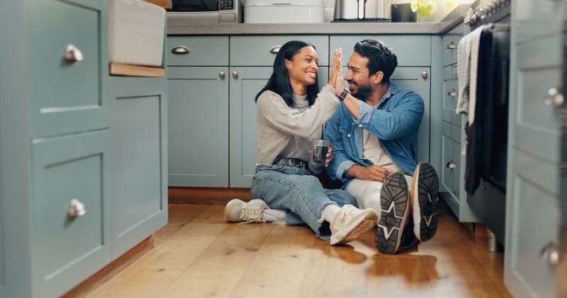 A happy couple high-fiving on the kitchen floor in a modern home, symbolizing positive decision-making about renting or buying their home.