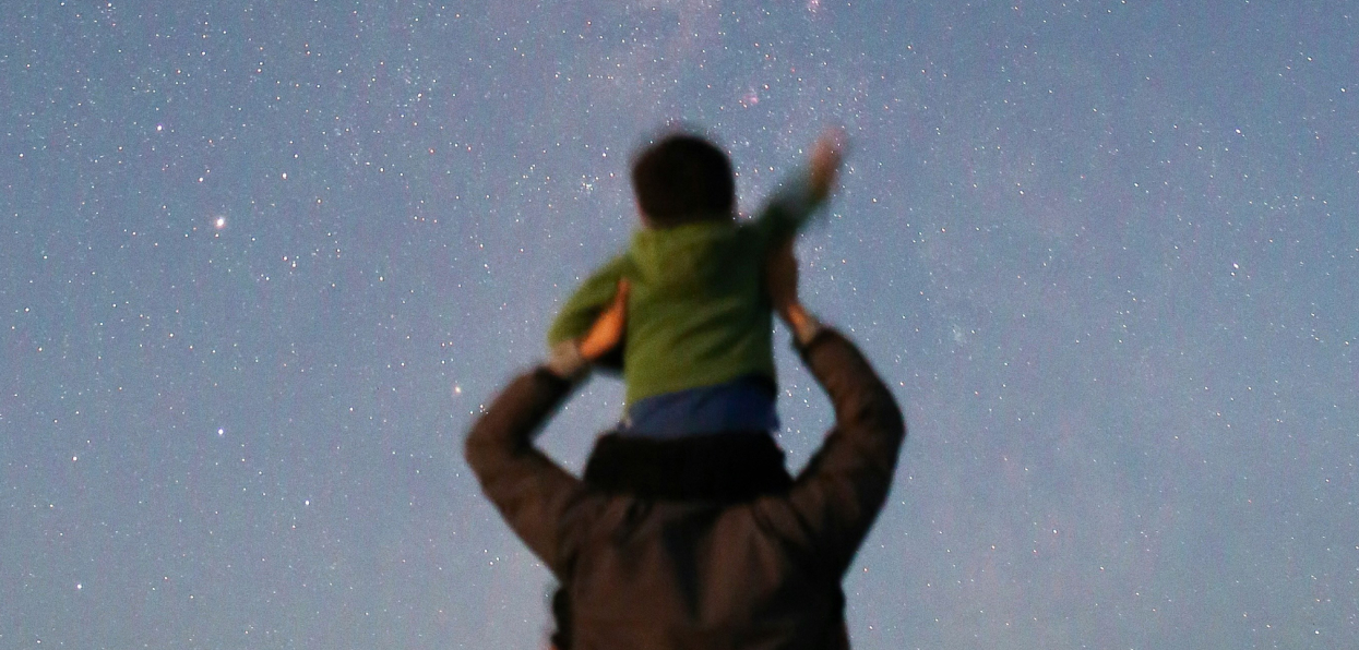 Rear-view of a child sitting on parent's shoulders pointing at the night sky