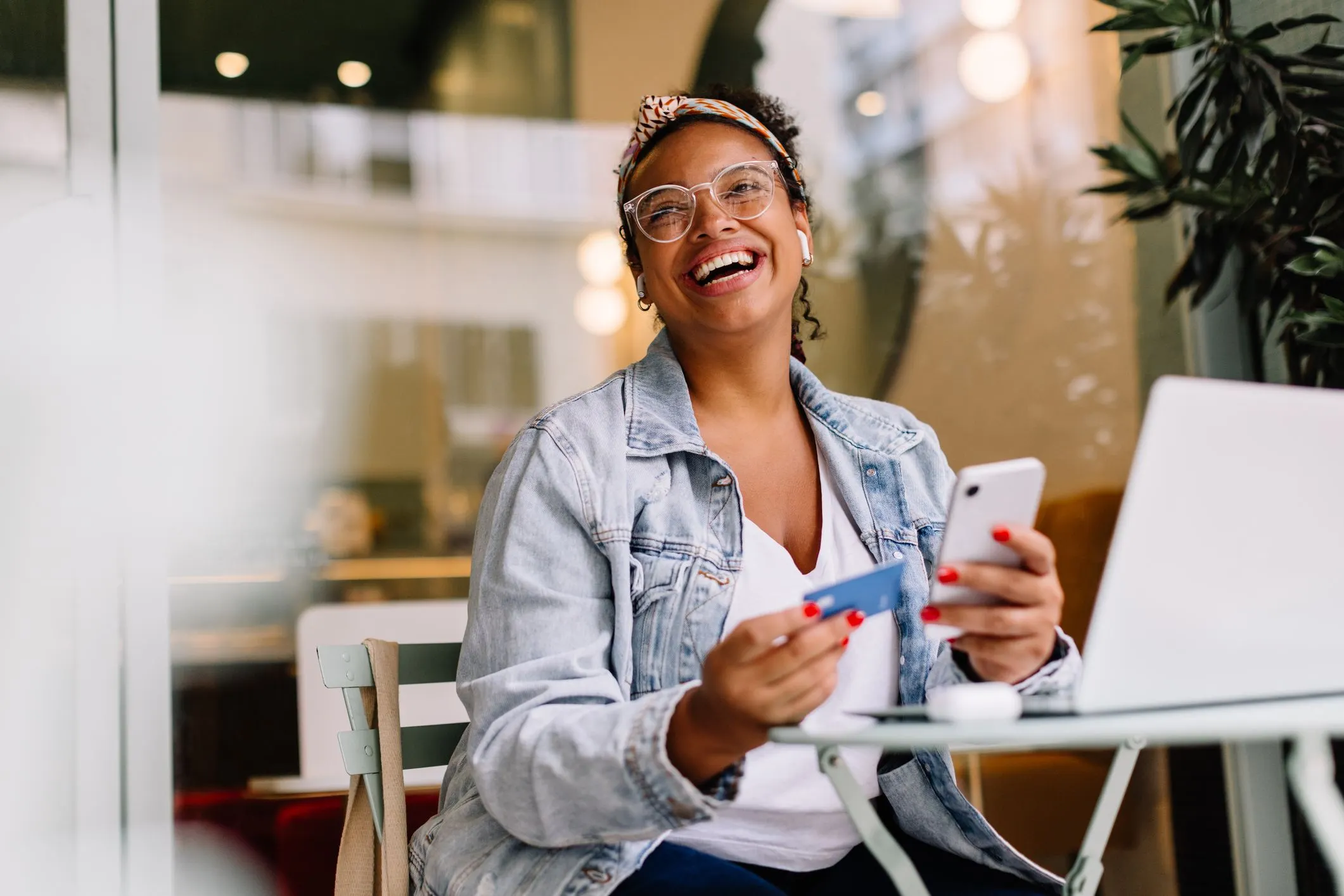 A cheerful woman sitting outdoors at a café, holding a smartphone in one hand and a credit card in the other.